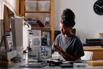Black female technician sitting at table in workshop and repairing system unit using screwdriver - obrazy, fototapety, plakaty