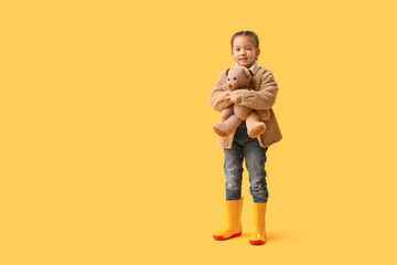 Little Asian girl in rubber boots with toy bear on yellow background