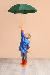 Little boy in rubber boots with umbrella near beige wall