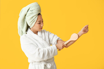 Young Asian woman in bathrobe with massage brush after shower on yellow background
