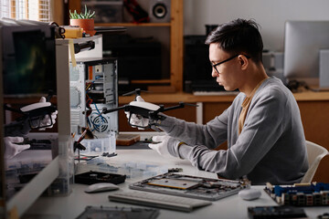 Young caucasian electronic repairman attentively examining quadcopter sitting at desk in workshop