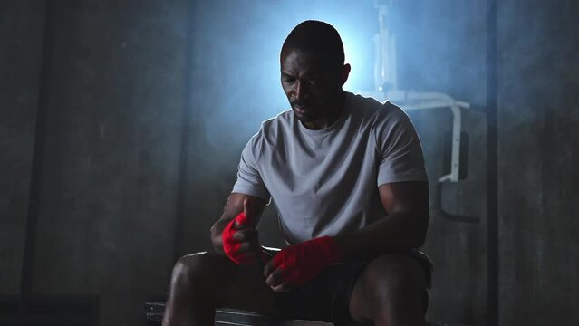 Fitness workout in gym. African American man fighter preparing for fight wrapping hands with red boxing wraps sports protective bandages in gym. Strong man ready for fight boxing sparring training