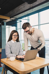 Colleagues communicate while working with a laptop in the office. Two workers in a modern workspace. Two young businessmen are sitting together at a table.