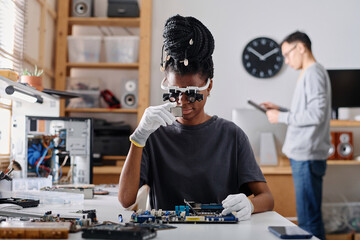 Female technician wearing glasses with loupes examining CPU, her male coworker standing on...