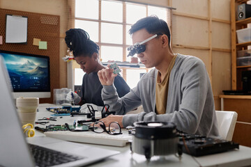 Young repairman in glasses with loupes examining CPU sitting next to his black female colleague - Powered by Adobe
