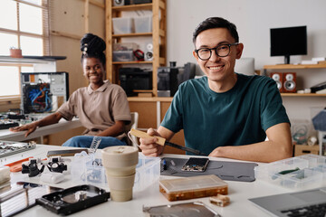 Caucasian technician smiling at camera while soldering cellphone, his black coworker sitting on...