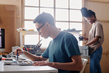 Young man repairing PC element with soldering iron, his black coworker standing on background