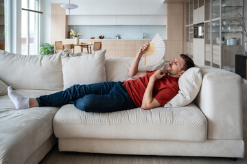 Overheated perspiring man lying on sofa, waving with hand fan, suffering from high temperature in...