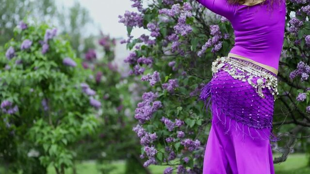 Woman in purple dress waves body during belly dance near bushes of blossom lilac. Slow motion