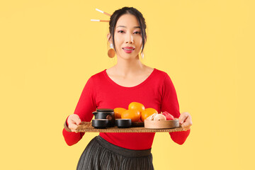 Young Asian woman holding tray with tea, mandarins and fortune cookies on yellow background....