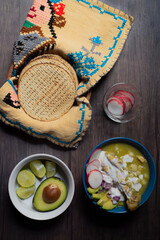 Comida Mexicana. Mesa de madera, puesta con plato lleno de pozole verde, con tostadas rabanos y aguacate. Todo entra en ecncuadre, vertical.