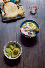 Comida Mexicana. Plato de Pozole Verde con rabanos, aguacate y tostadas en tortillero, sobre mesa de madera. Vertical.