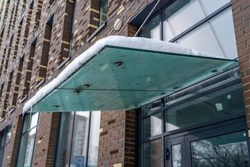 Fresh snow accumulates on the glass awning of an urban building, showcasing serene beauty of a city under winters gentle embrace.