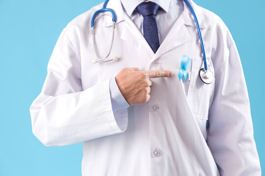Male doctor pointing at blue ribbon on color background, closeup. Prostate cancer awareness concept