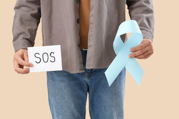 Young man holding paper with word SOS and blue ribbon on beige background, closeup. Prostate cancer...