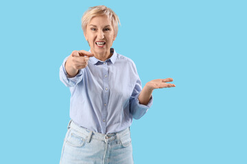 Angry mature woman pointing at viewer on blue background. Accusation concept
