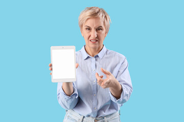 Angry mature woman with tablet computer pointing at viewer on blue background. Accusation concept