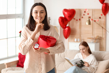 Young lesbian woman with gift and engagement ring showing silence gesture at home on Valentine's Day