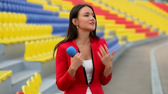 Woman in red jacket holds microphone and speaks near seats on sports stadium