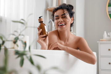 Young African-American woman with bottle of cream taking bath at home