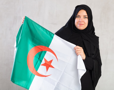 calm Muslim woman in traditional black hijab holds flag of Algeria. Portrait of female Muslim with Algerian flag on gray background