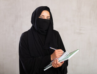young calm girl with covered face by black burka holds several notebooks in her hands and writes...