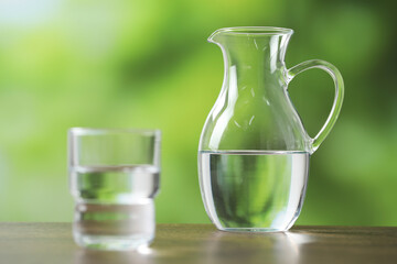 Jug and glass with clear water on wooden table against blurred green background, selective focus