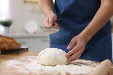 Making bread. Man sprinkling flour onto dough at wooden table in kitchen, closeup