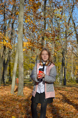 beautiful girl has fun while walking through the autumn park and fallen maple leaves. holding a glass of hot drink. the child smiles, laughs and enjoys the warmth and fresh air