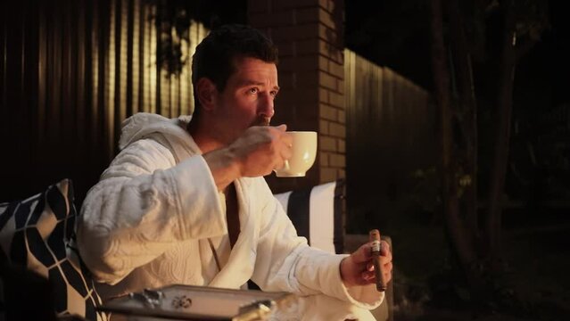 Man in bathrobe holds cigar and drinks coffee near burning fire on porch. Slow motion