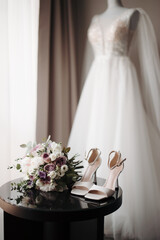 Modern accessories of the bride. Bouquet and high-heeled shoes on a black table and wedding dress on a mannequin in a hotel room near the window