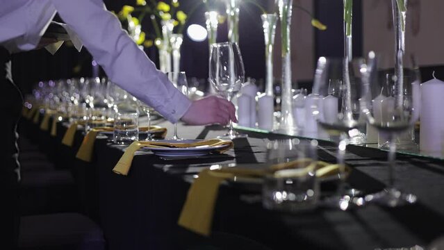 Banquet table for dinner with candles and yellow flowers in tall vases getting set by unknown butler in formal clothes. Luxury decorated restaurant ready for accepting anniversary celebration event.