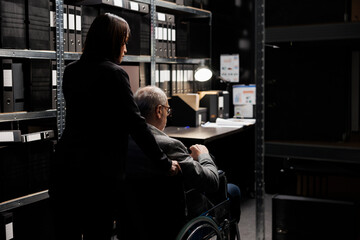 Private detective with disability in wheelchair and assisting coworker in investigation agency file cabinet storage office surrounded by criminology cases folders on cabinet shelves