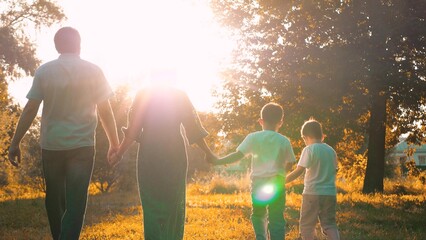 Joyful mother and father with sons stroll hand in hand across meadow during family weekend. Small...