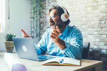 Cheerful Italian freelance man singing and listening to music with headphones while working with...