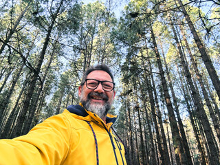 Cheerful adult happy man taking selfie picture in the woods enjoying outdoors leisure activity in national forestal park alone. Adventure and lifestyle people. High trees in background. Sustainability