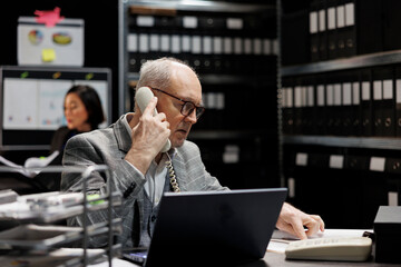 Accounting elderly businessman offering consultancy during phone call in bookkeeping workplace. Professional senior employee in archival depository filled with document folders and flowcharts - obrazy, fototapety, plakaty
