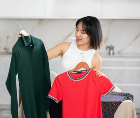 Glad young Asian woman looking cheerfully at dresses held in hand for choice