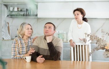 Family members quarreling in the kitchen and old woman trying to calm angry man sitting at the table