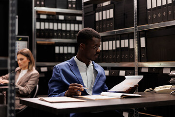 African american bookkeeper analyzing management files, searching for administrative report in corporate repository. Businessman working late at night at bookkeeping record in storage room