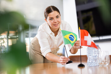 Positive young woman putting little flag of Brazil on the table with flag of Switzerland in...