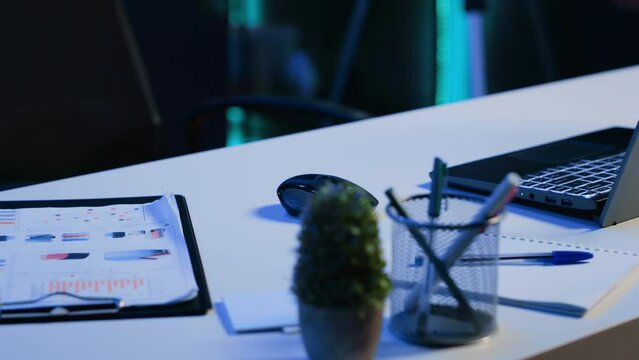 Office desk filled with productivity items in empty home interior illuminated with neon lights at night. Laptop on table and other work related objects in apartment room, camera A close up shot