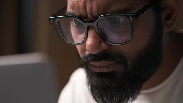 Close-up portrait of a concentrated and thoughtful indian man in glasses working on a laptop, thinking, doing online learning, contemplating, pondering.