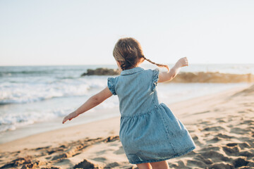 girl with braids in a blue denim dress twirling in the sand at the beach with her back to the camera