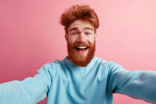 Ecstatic ginger bearded guy in blue sweatshirt takes selfie isolated on pink background