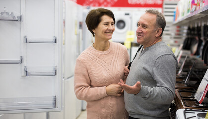 Middle aged wife and husband picking new refrigerator in shop of household appliances.