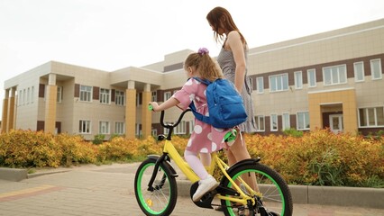 Playful little girl pupil riding on bike at schoolyard with mother arriving to elementary school outdoor. Adorable female kid child driving cycle with mom help learning approach campus primary lesson