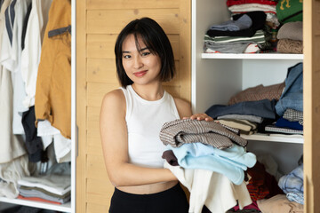 Happy young Asian girl holding some items of clothing standing in front of the wardrobe
