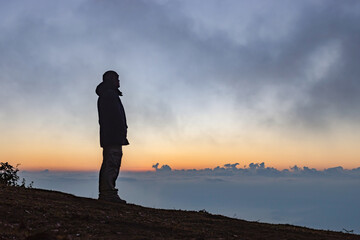 Young man standing alone on top mist mountain and staring at sunrise, cloudy sky.