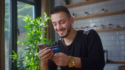 Smiling man holding credit card and smartphone enters credit card number to online purchases. Male using instant easy mobile payments, making purchase in online store